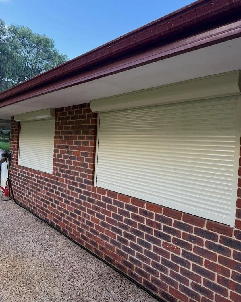 Exterior of a brick house with two windows covered by closed white roller shutters, and a brown roof overhang, on a cloudy day.