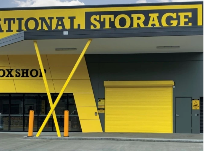 Exterior of a storage facility with a yellow roller shutter door, yellow and black signage, and yellow posts in front.