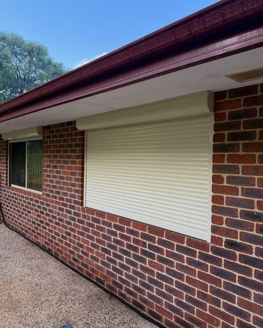 A brick house exterior with a window and a cream-colored closed roller shutter under a brown roof with a bright blue sky above.