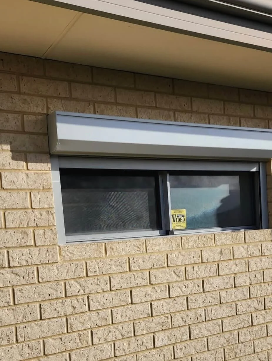 A horizontal rectangular window with sliding glass panes set in a beige brick wall, with a small yellow sticker on the right pane that says 'Video' and a warning.