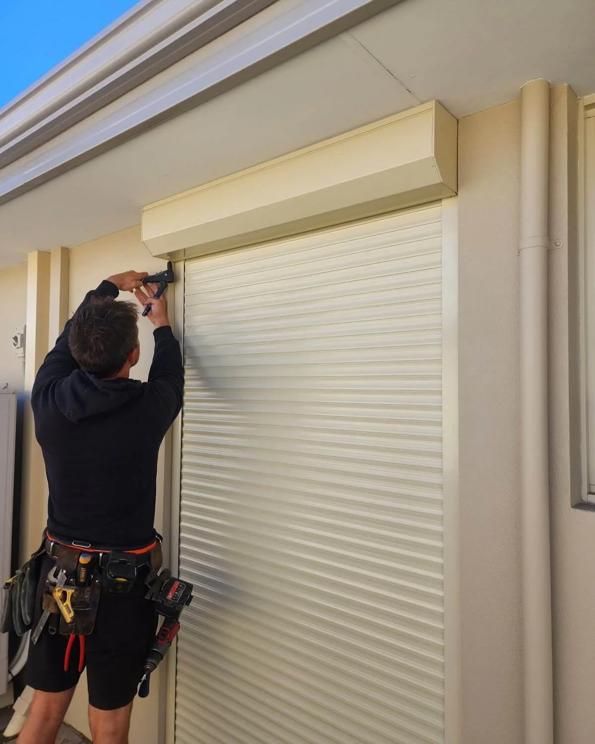 Nathan Russell from Coastal Shade and shutters is installing or repairing a beige rolling shutter door outside a building, using a power drill while wearing a tool belt.