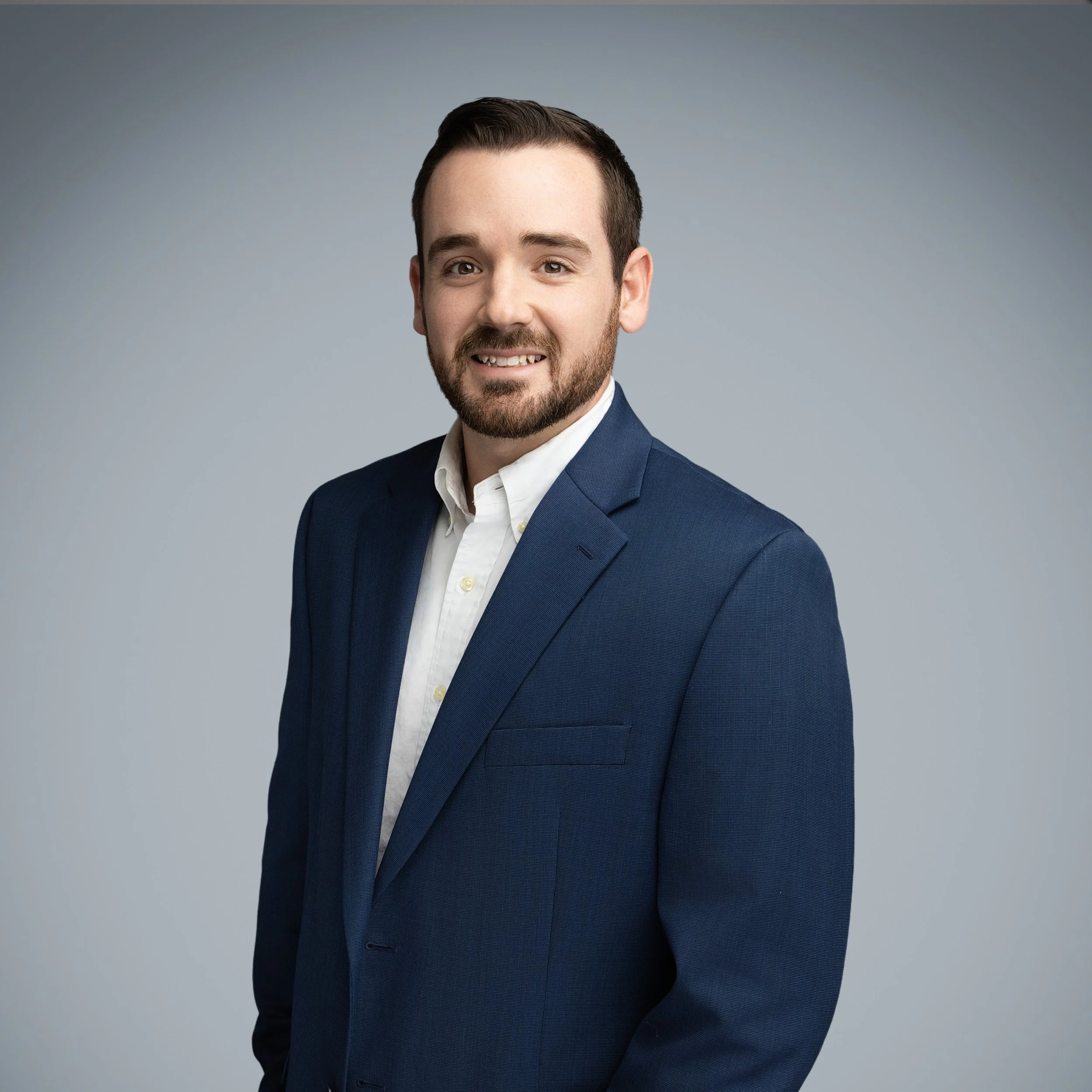 A young man in a navy blue suit with a white shirt, smiling, posed against a plain gray background.
