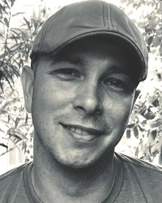 A smiling man wearing a baseball cap outdoors, with leafy background.