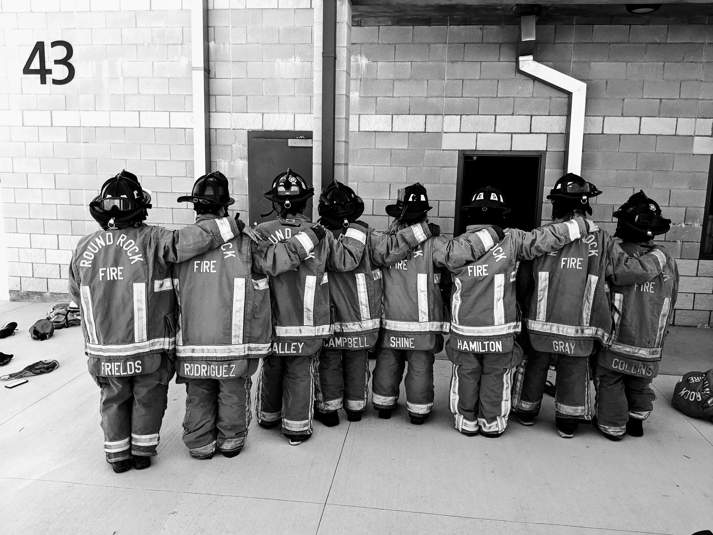 A group of nine firefighters in turnout gear and helmets standing outside a building, with arms around each other's shoulders, facing a wall.
