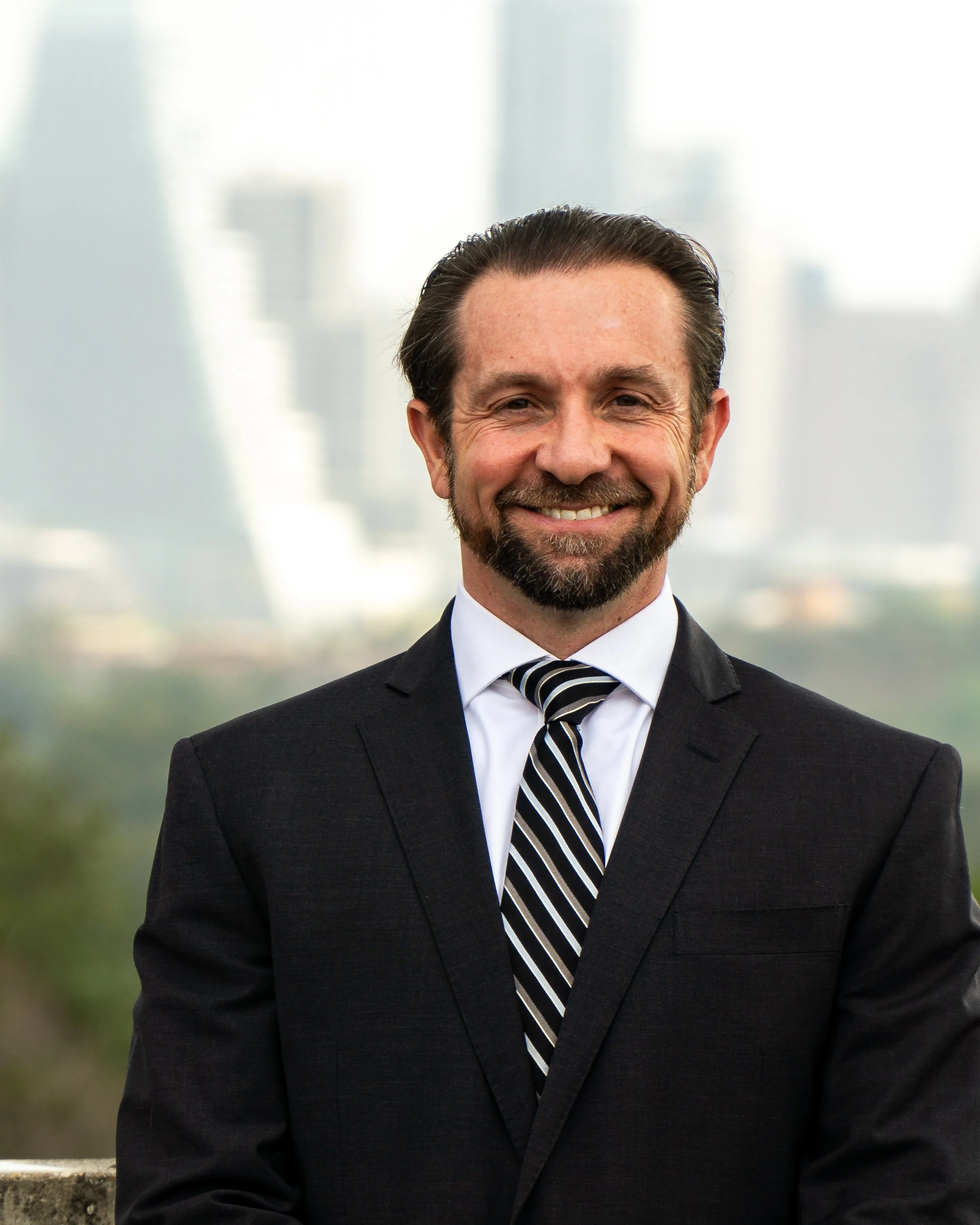 A professional man in a black suit, white shirt, and striped tie smiling outdoors with a city skyline in the background.