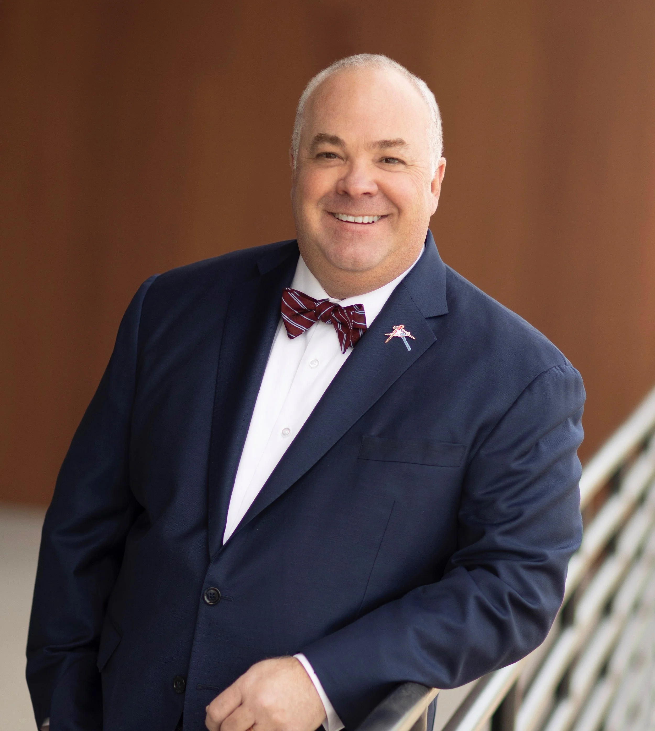 Man in a navy suit with a striped bow tie, smiling, standing indoors with a brown background and staircase.
