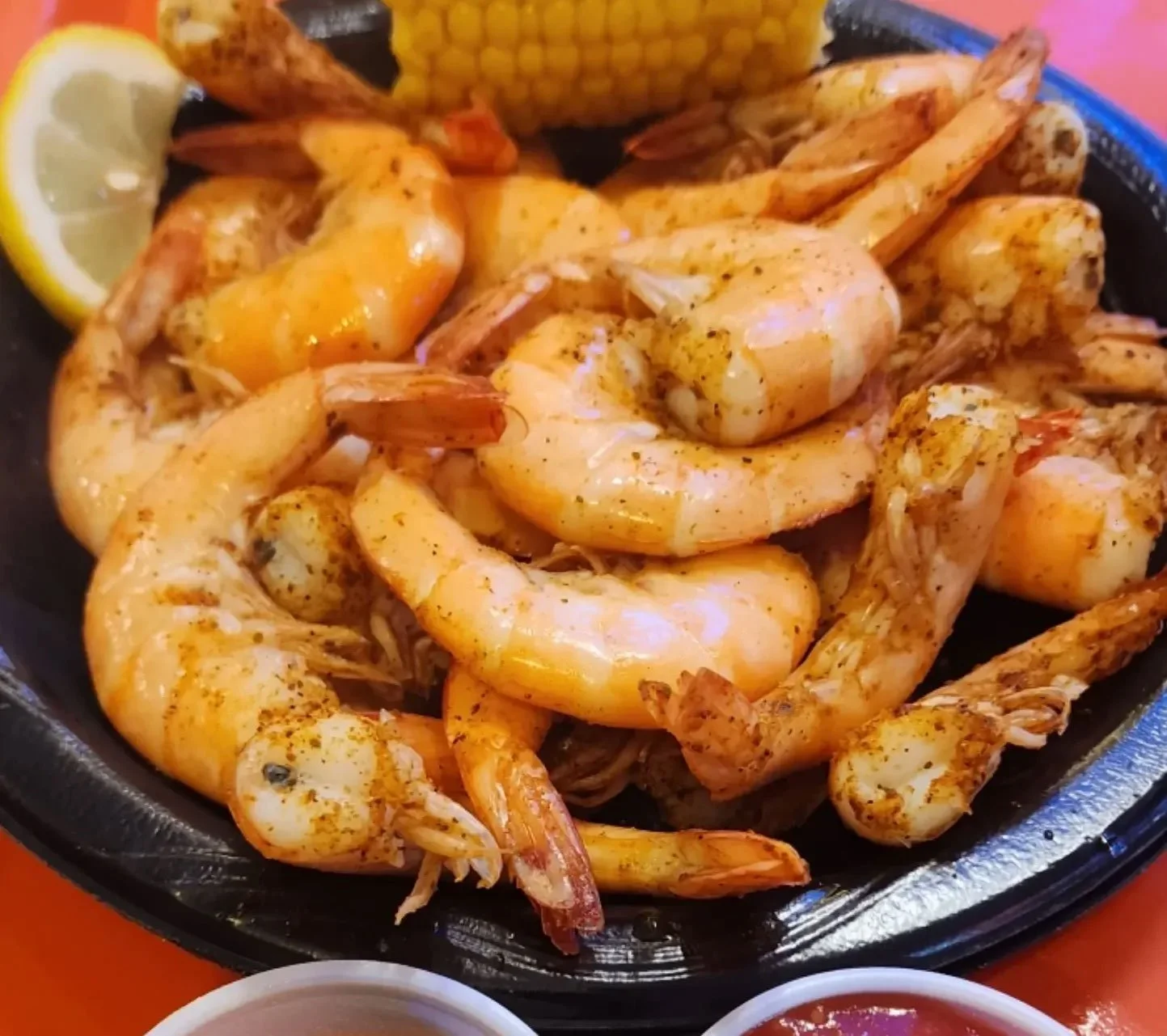 A bowl of cooked shrimp seasoned with spices, with lemon wedge and corn on the cob in the background, served on a black plate.