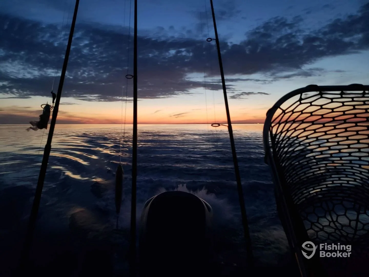 View from a boat at sunset showing fishing rods and a fishing net against a colorful sky with clouds over calm ocean water.