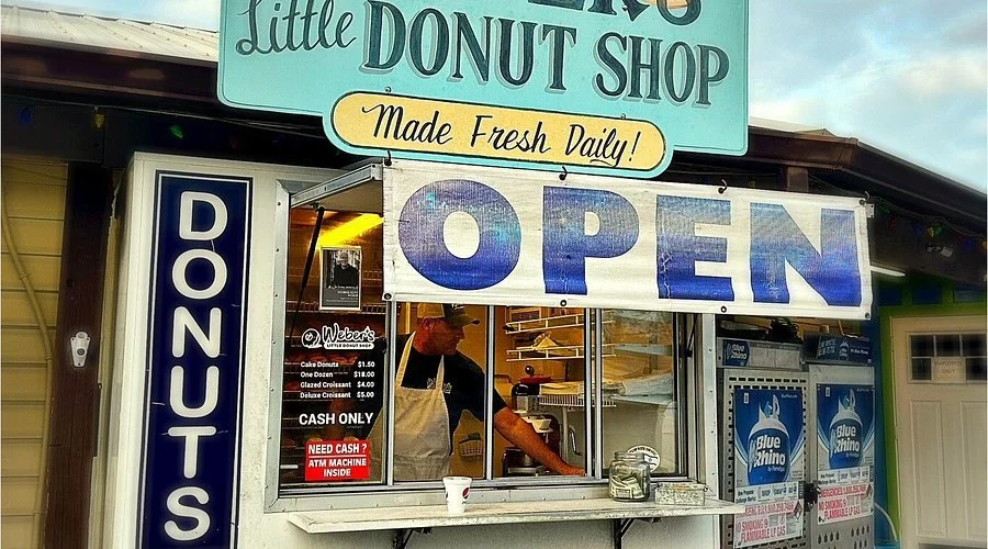 Small donut shop with a large illuminated 'OPEN' sign and a vertical sign listing donut varieties and prices. There is a man inside preparing food, and vending machines outside.
