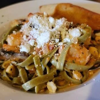 A plate of green pasta with shrimp, mushrooms, and grated cheese, served with a slice of garlic bread.