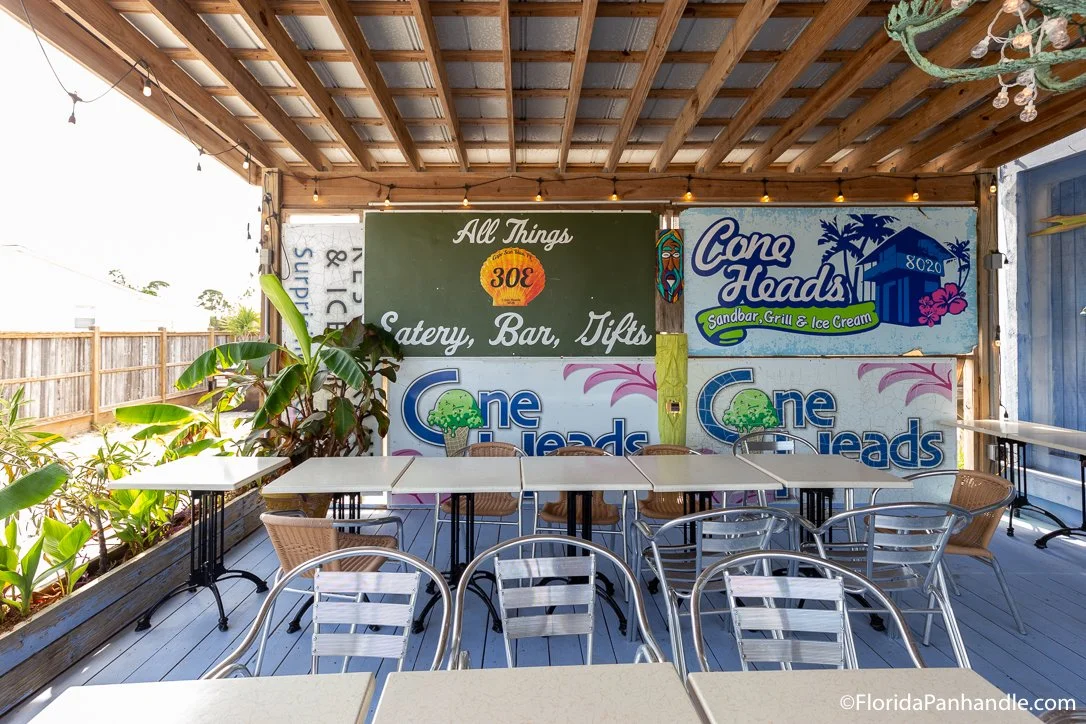 Covered outdoor seating area at a beachside café with tables and chairs, signs advertising food and drinks, and tropical plants.