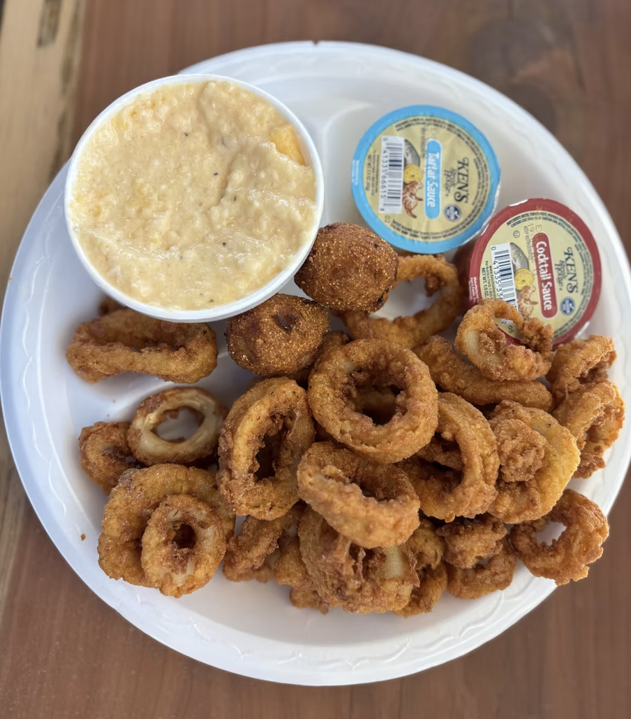 A white disposable plate filled with fried calamari rings, two fried hush puppies, a cup of tartar sauce, and a cup of cocktail sauce.