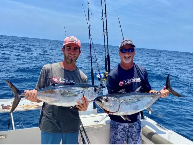 Two men on a boat holding large fish they caught, with fishing rods in the background, out in the ocean on a sunny day.