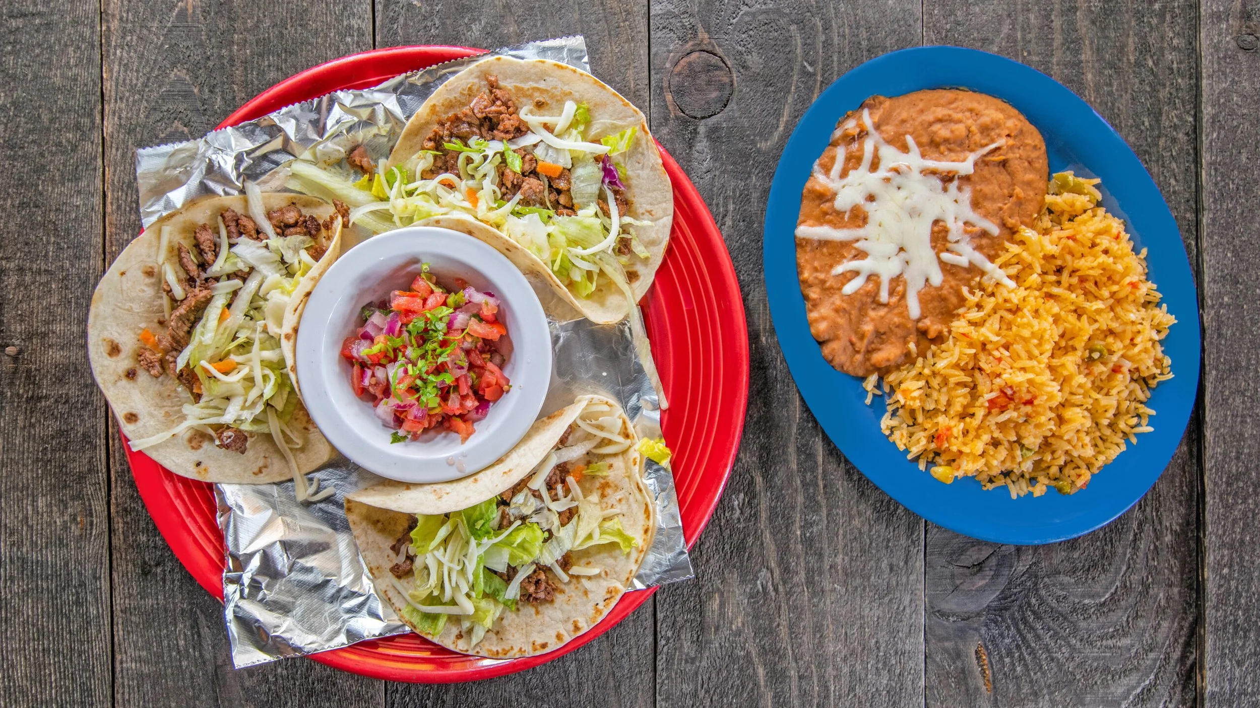 A plate of beef tacos with shredded lettuce, diced tomatoes, and chopped onions served with a side of rice and beans topped with cheese.