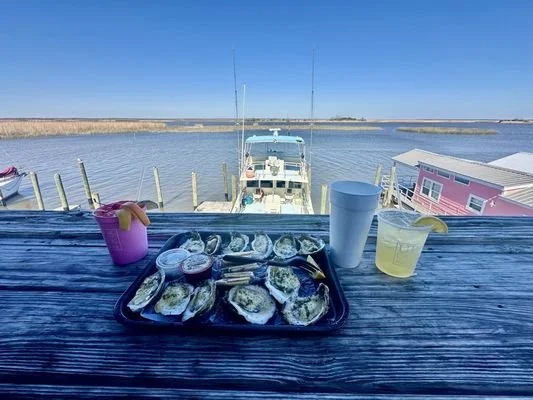 A tray of oysters on a wooden table overlooking a marina with boats and a clear blue sky.