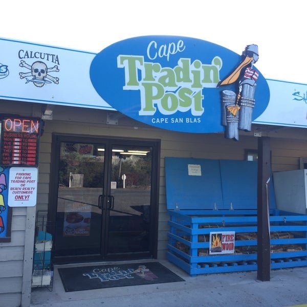 Entrance of a store called Cape Tradin Post in Cape San Blas, Florida, with a large blue sign featuring a pirate figure and a skull and crossbones. There are several blue crates outside, one marked with a firewood sign, and a neon open sign in the window.