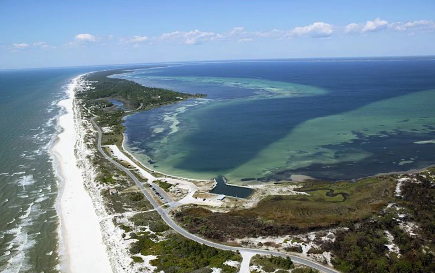 Aerial view of a coastline with sandy beach, lush green land, and a large body of water separated by a dam or barrier, with clear blue sky and clouds overhead.