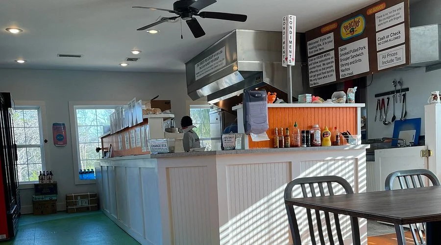 Inside a small restaurant or fast-food counter with a white counter, condiment bottles, a digital order display, and a person working behind the counter. There are windows letting in natural light, and seating with metal chairs visible in the foreground.