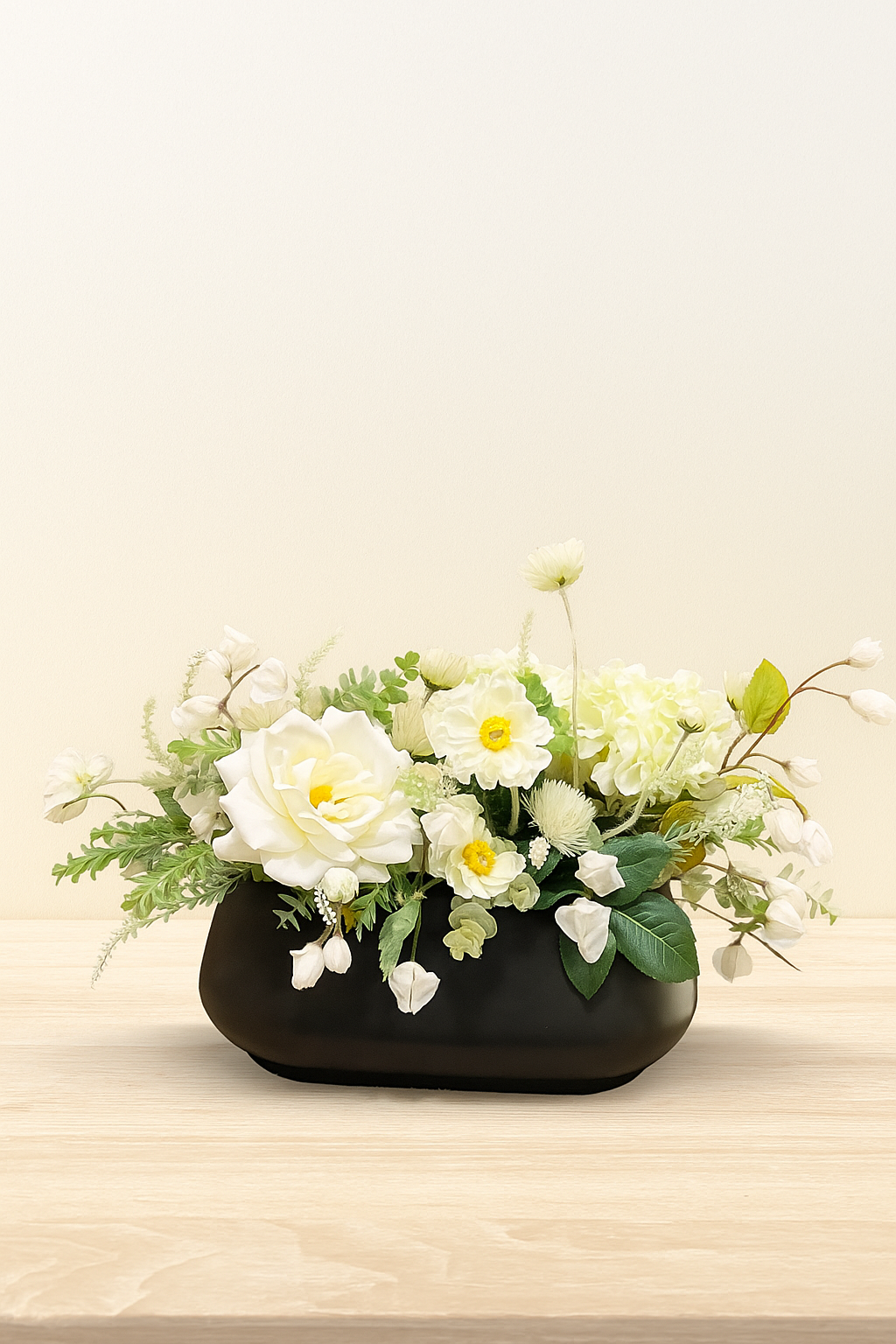 White flower arrangement in a black vase on a light wood surface against a plain white background.
