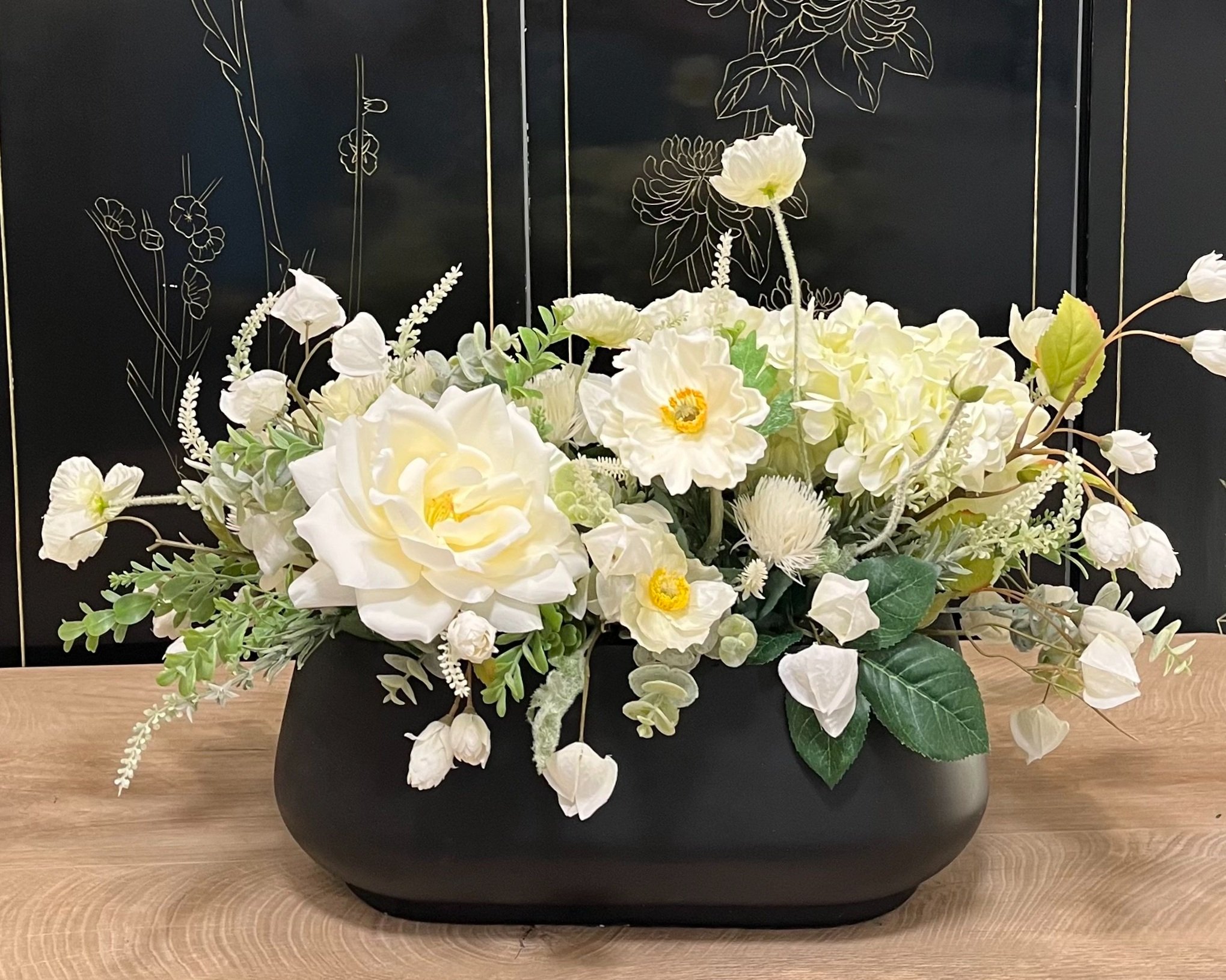 White floral arrangement in a black vase on a wooden surface with a black background featuring white line drawings of flowers.