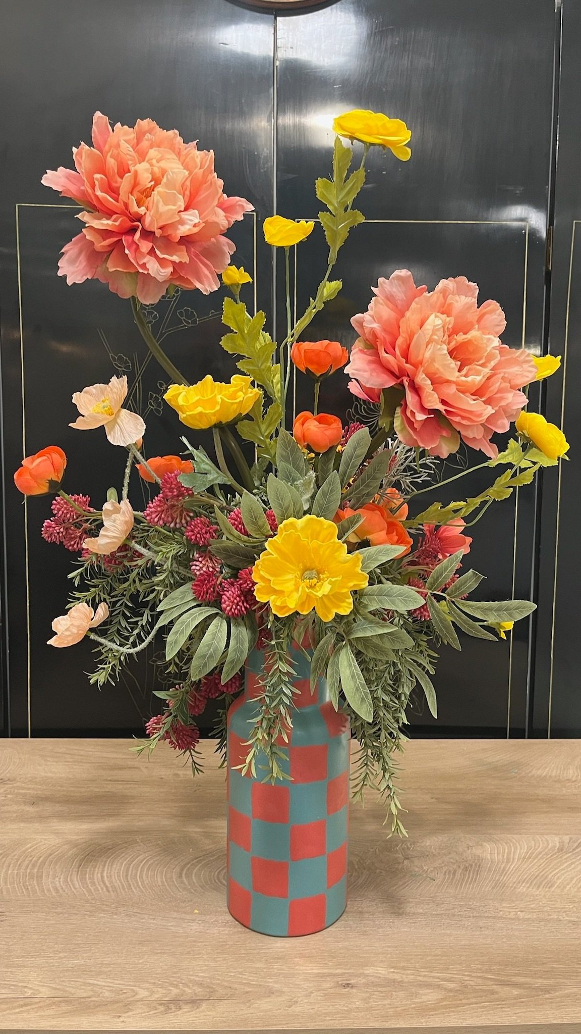 A colorful flower arrangement in a tall vase with a checkered pattern. The flowers include large pink blossoms, yellow flowers, orange blooms, and various green foliage, set against a dark backdrop with a wooden surface.