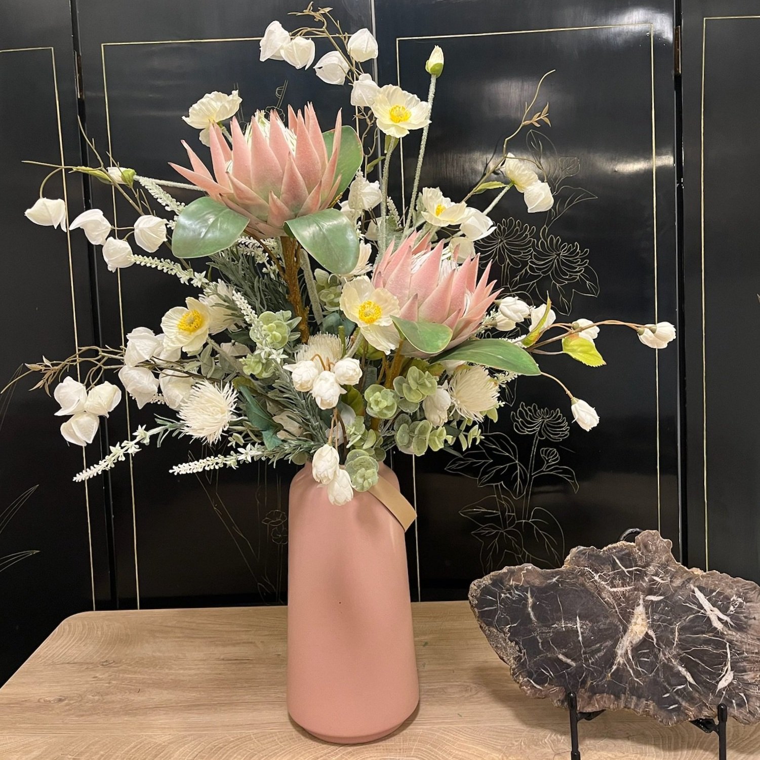 A pink vase with a large floral arrangement of pink protea, white flowers, and green foliage on a wooden table, with a black decorative background and a dark stone sculpture nearby.