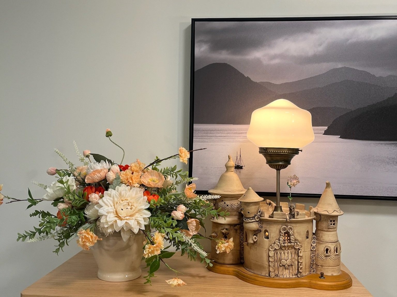 Decorative table with a floral arrangement in a white vase, an ornate castle-shaped lamp with a lampshade, and framed landscape photo of mountains and water in the background.
