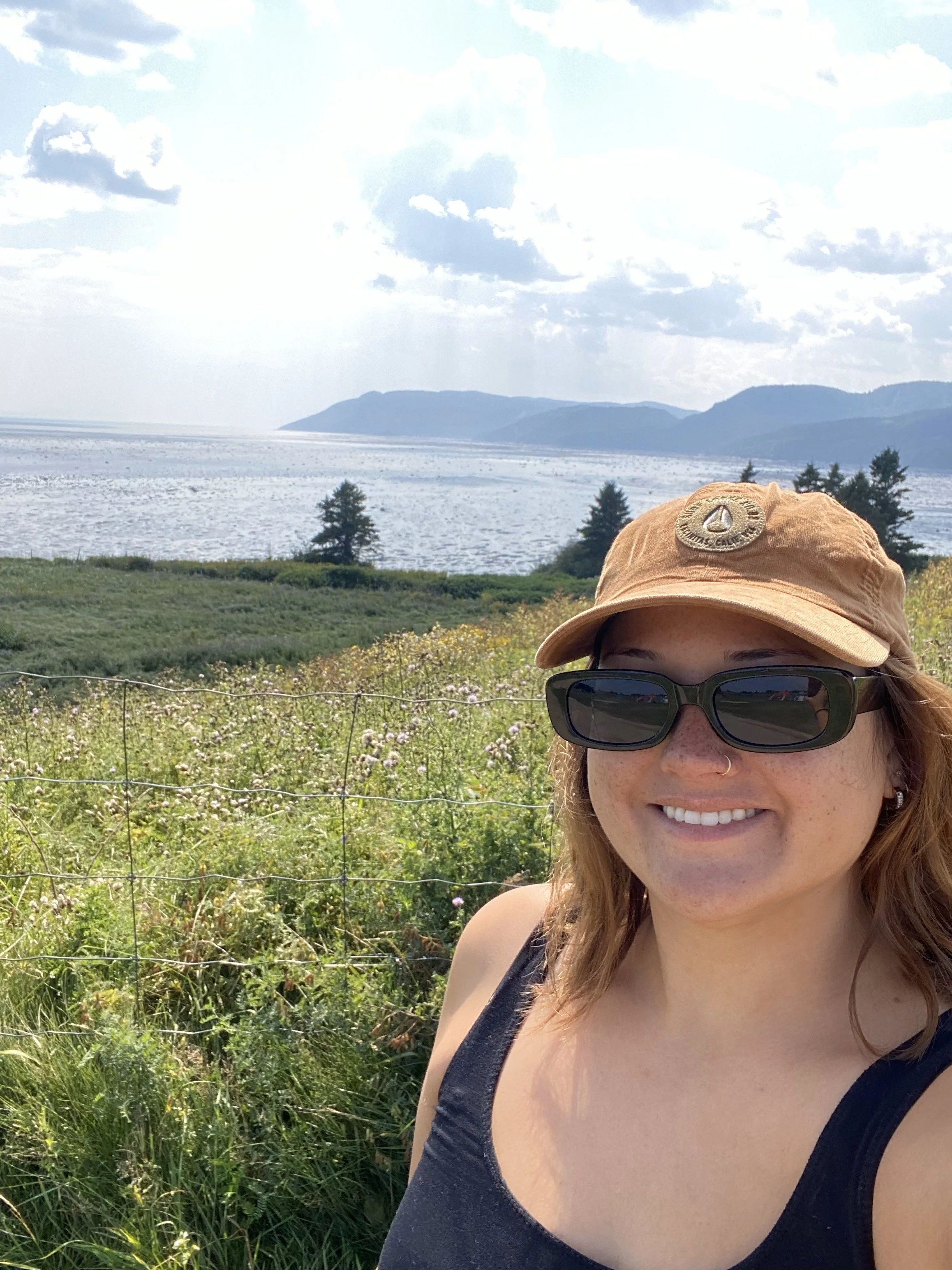 A young woman taking a selfie outdoors with mountains, a body of water, and a partly cloudy sky in the background. She is wearing a tan cap, black sunglasses, and a black tank top, and is smiling.