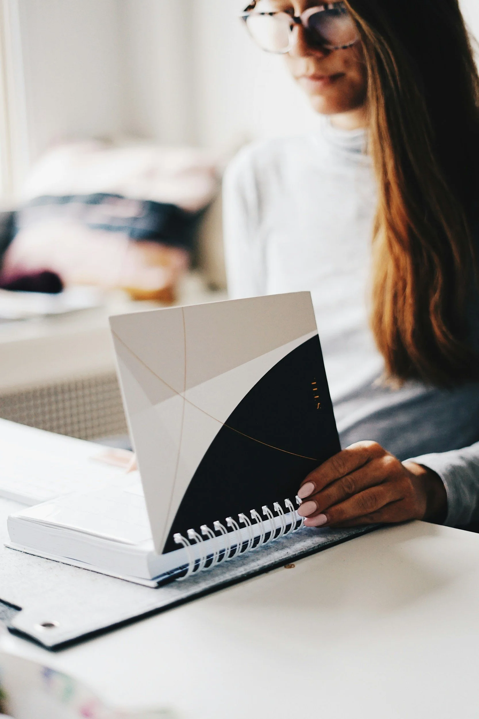 A woman with glasses reading a spiral-bound notebook with a black and white geometric cover, sitting at a white desk.