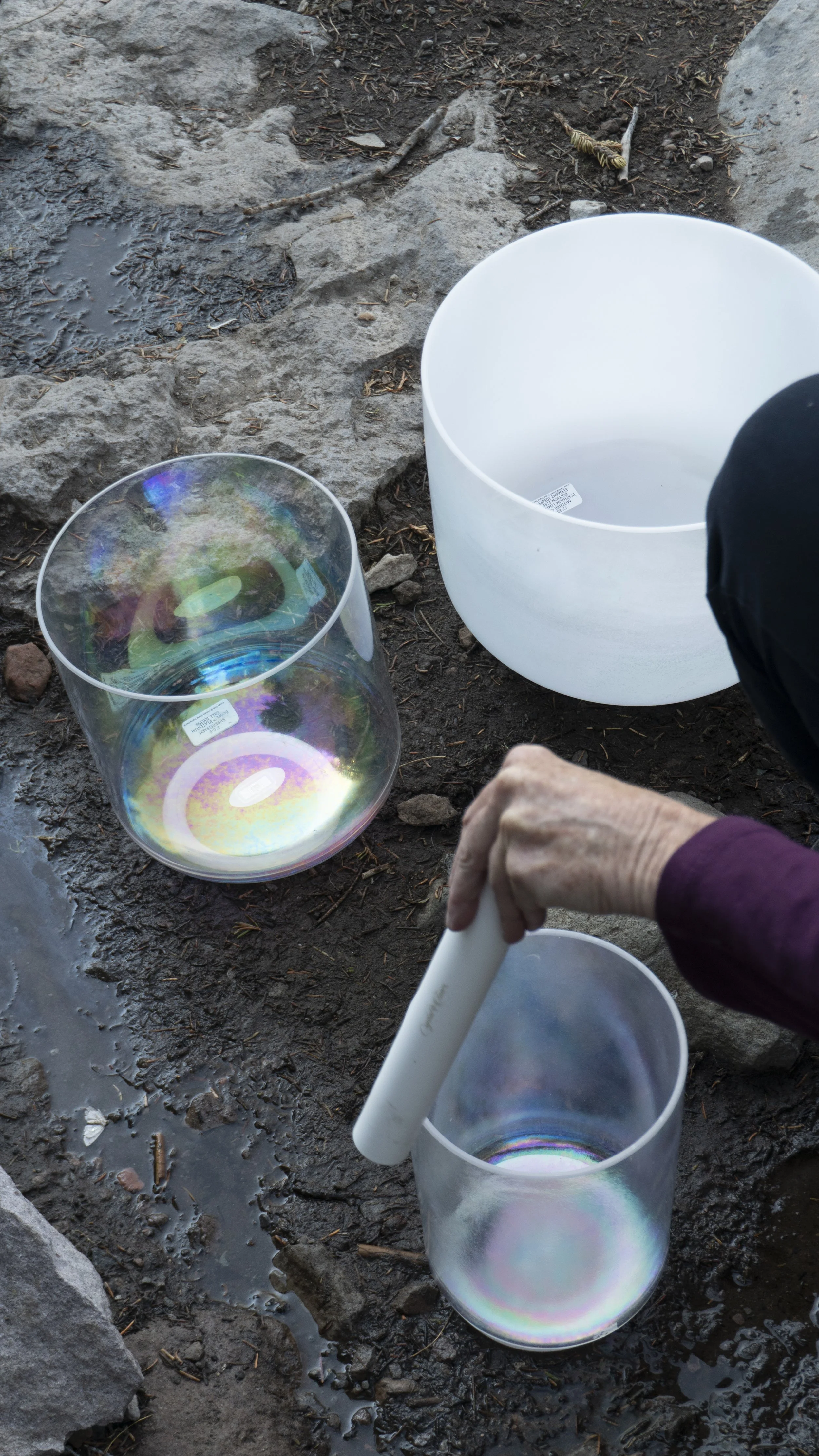 A person is holding a mallet about to strike a glass singing bowl outdoors, with two other larger singing bowls placed on wet, rocky ground nearby.