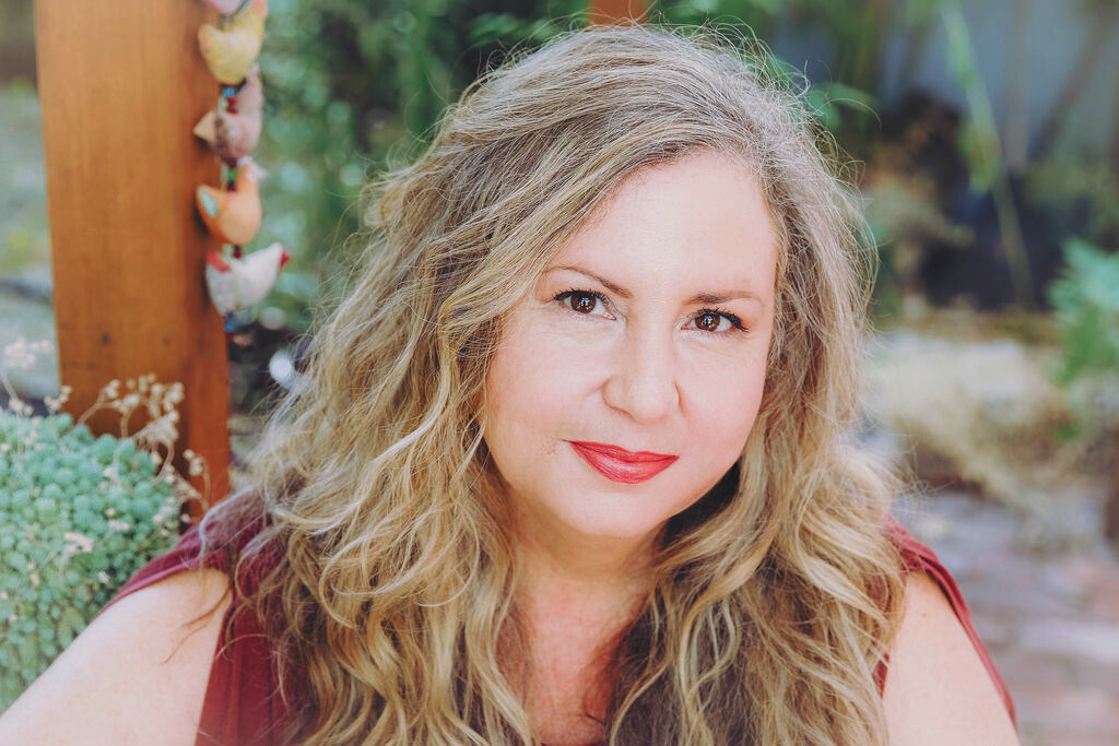 A woman with curly blonde hair and red lipstick looking at the camera, outdoors in a garden setting.