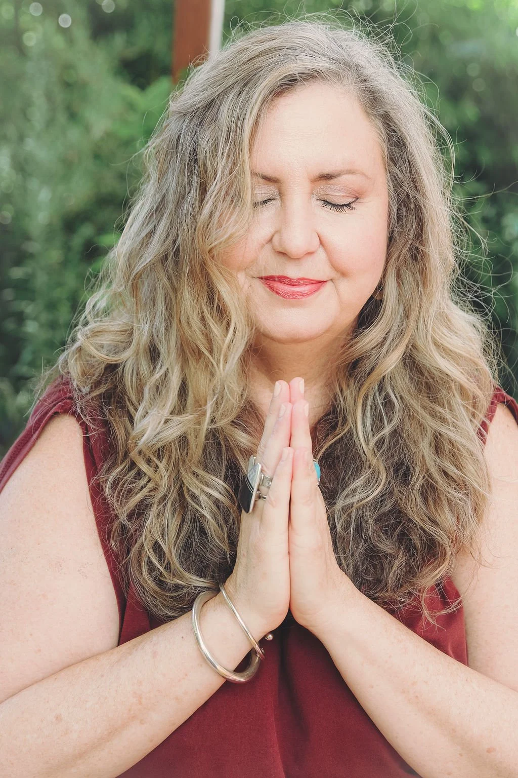 A woman with long, curly blond hair and closed eyes, dressed in a sleeveless maroon top, standing outdoors with a serene expression and hands pressed together in a prayer position.