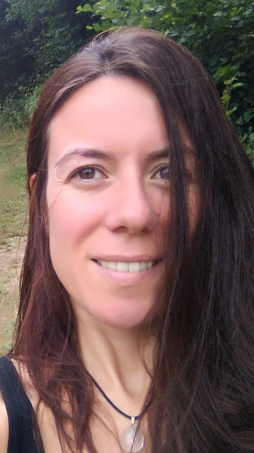 Close-up of a woman with long brown hair outdoors, smiling, with a green leafy background.