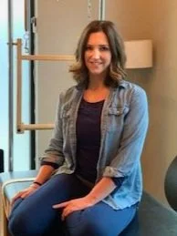 A woman with shoulder-length brown hair sitting on a blue chair in an indoor space, smiling, with a lamp and wooden rack in the background.