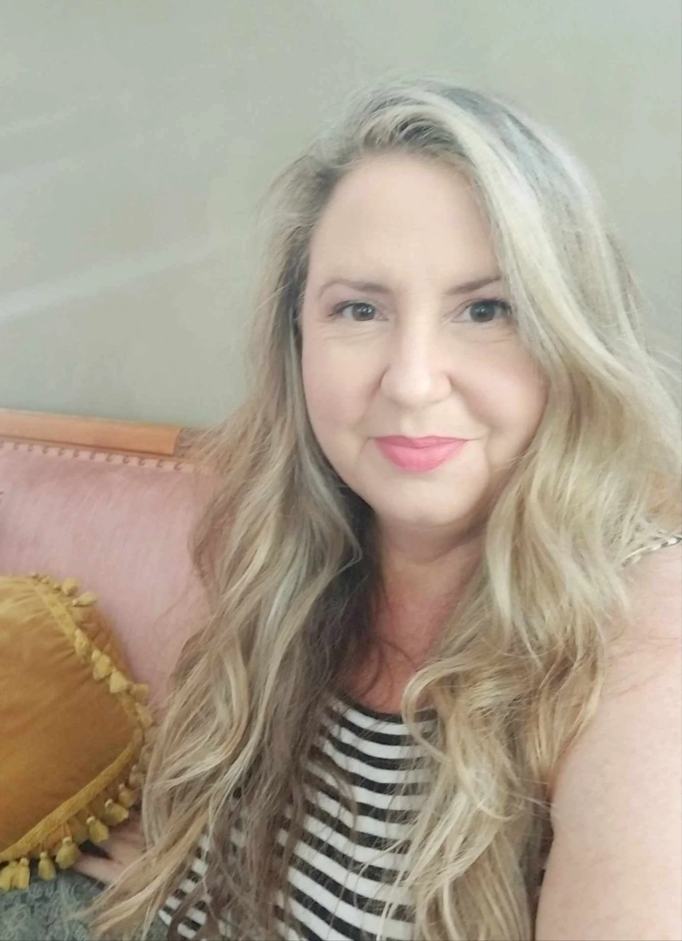 A woman with long, wavy blonde hair smiling indoors, wearing a striped top, sitting on a pinkish-beige couch with a yellow pillow.