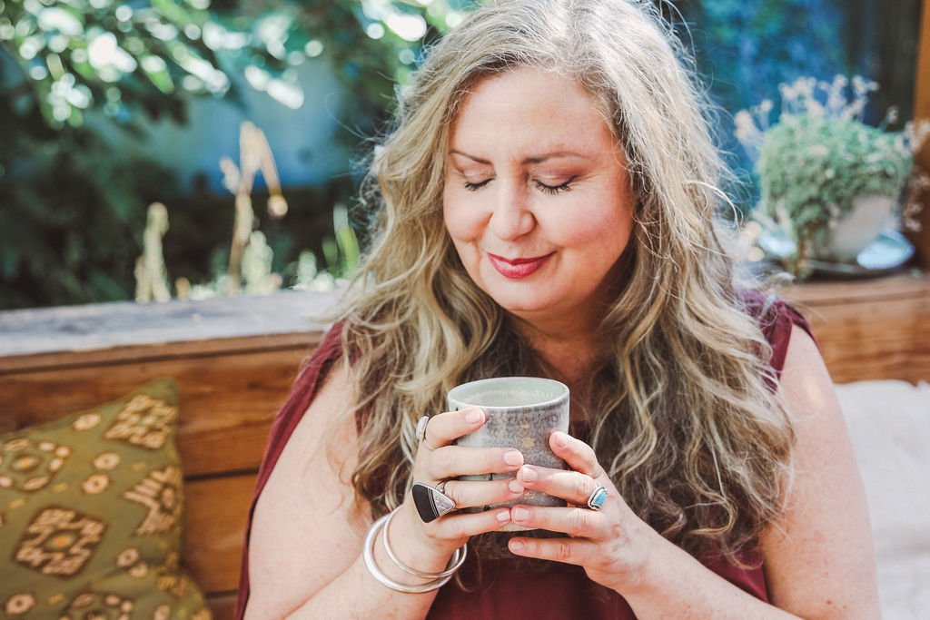 A woman with blonde curly hair and a content expression is holding a gray mug with both hands, eyes closed, sitting in a cozy outdoor setting with greenery and a wooden backdrop.