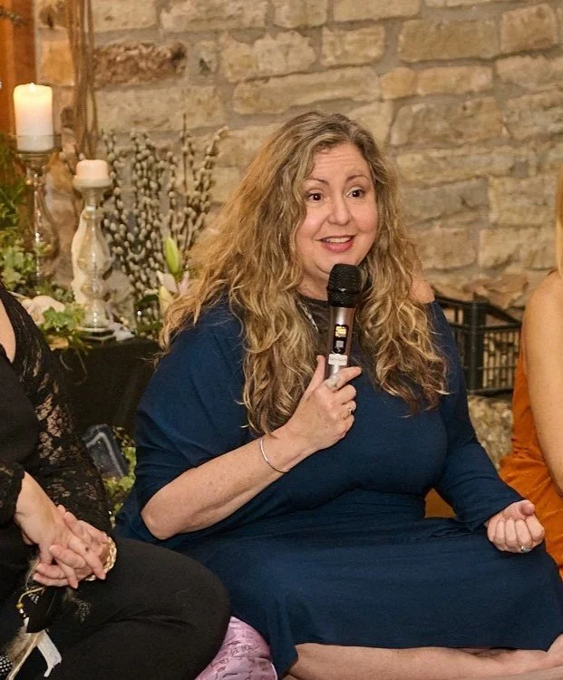 A woman with long curly blonde hair, wearing a blue dress, sitting and speaking into a microphone at an indoor gathering with a brick wall and candles in the background.