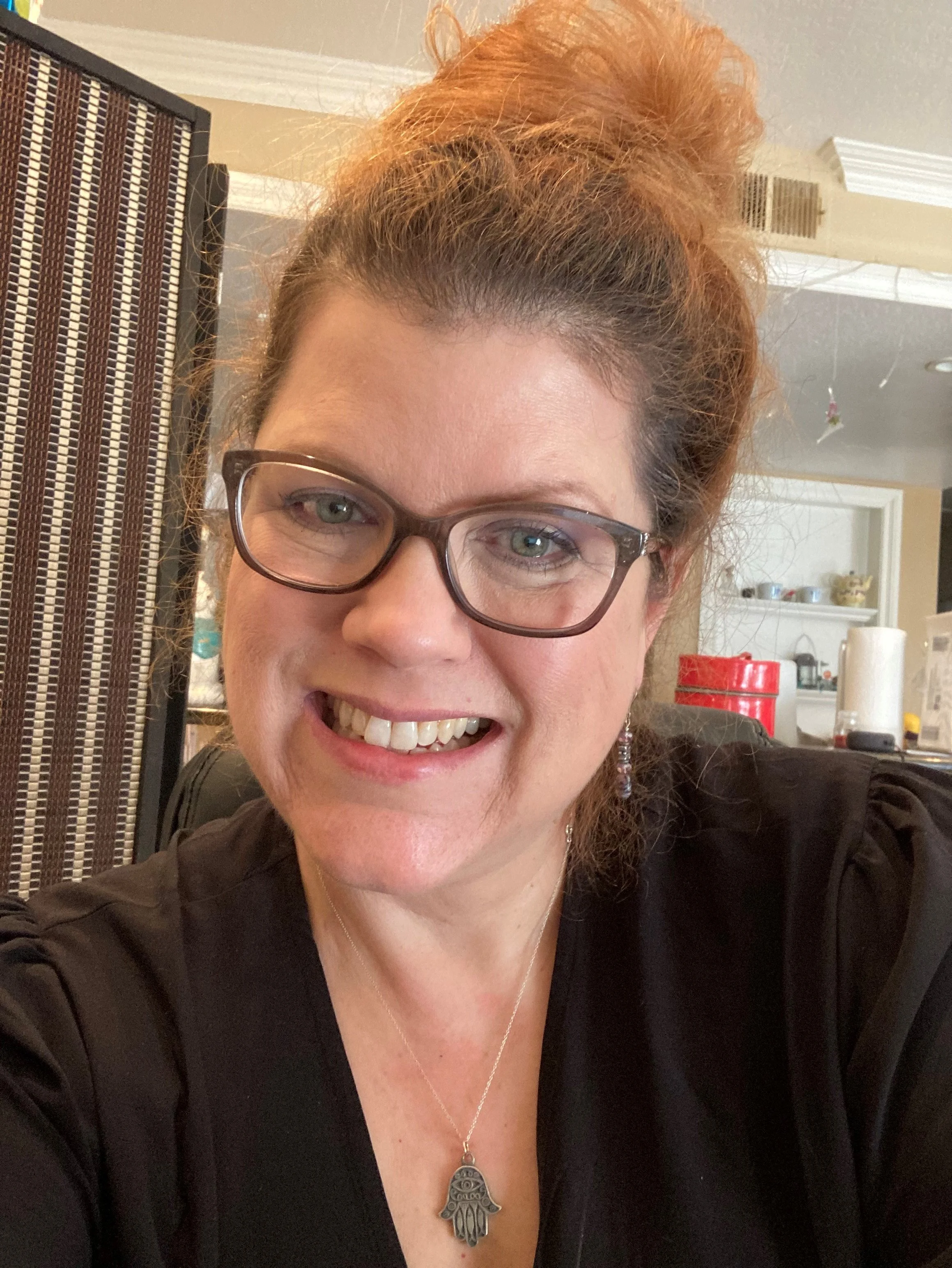 A woman with curly red hair styled in a high bun, wearing glasses and a black top, smiling.