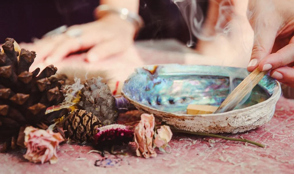 Person using a wooden stick to stir a soap mixture in an iridescent shell-shaped dish, surrounded by dried flowers and pinecones on a pink surface.