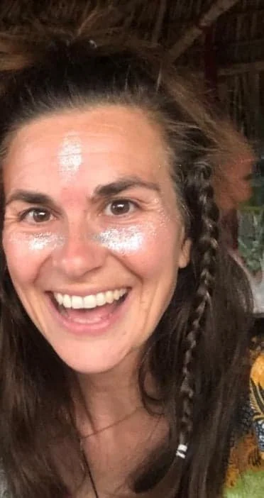 Woman smiling with face paint or sunscreen on forehead and cheeks, hair in braids, indoors with wooden ceiling and some greenery visible.