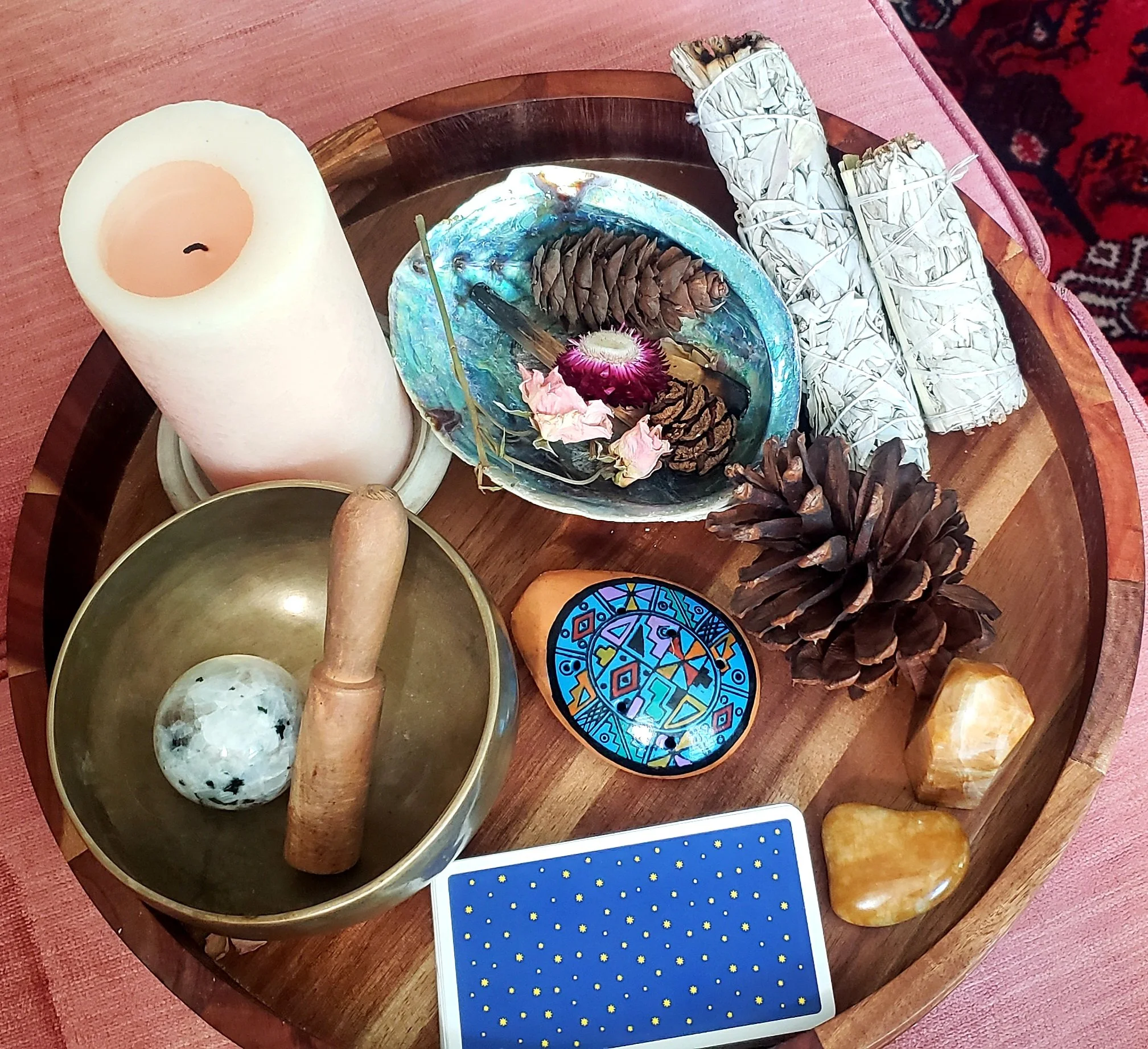 A wooden tray containing a white candle, a crystal ball, a decorative shell bowl with pinecones and flowers, two rolled bundles of sage tied with white string, tarot cards, and a large amber-colored stone, set on a pink cloth surface.