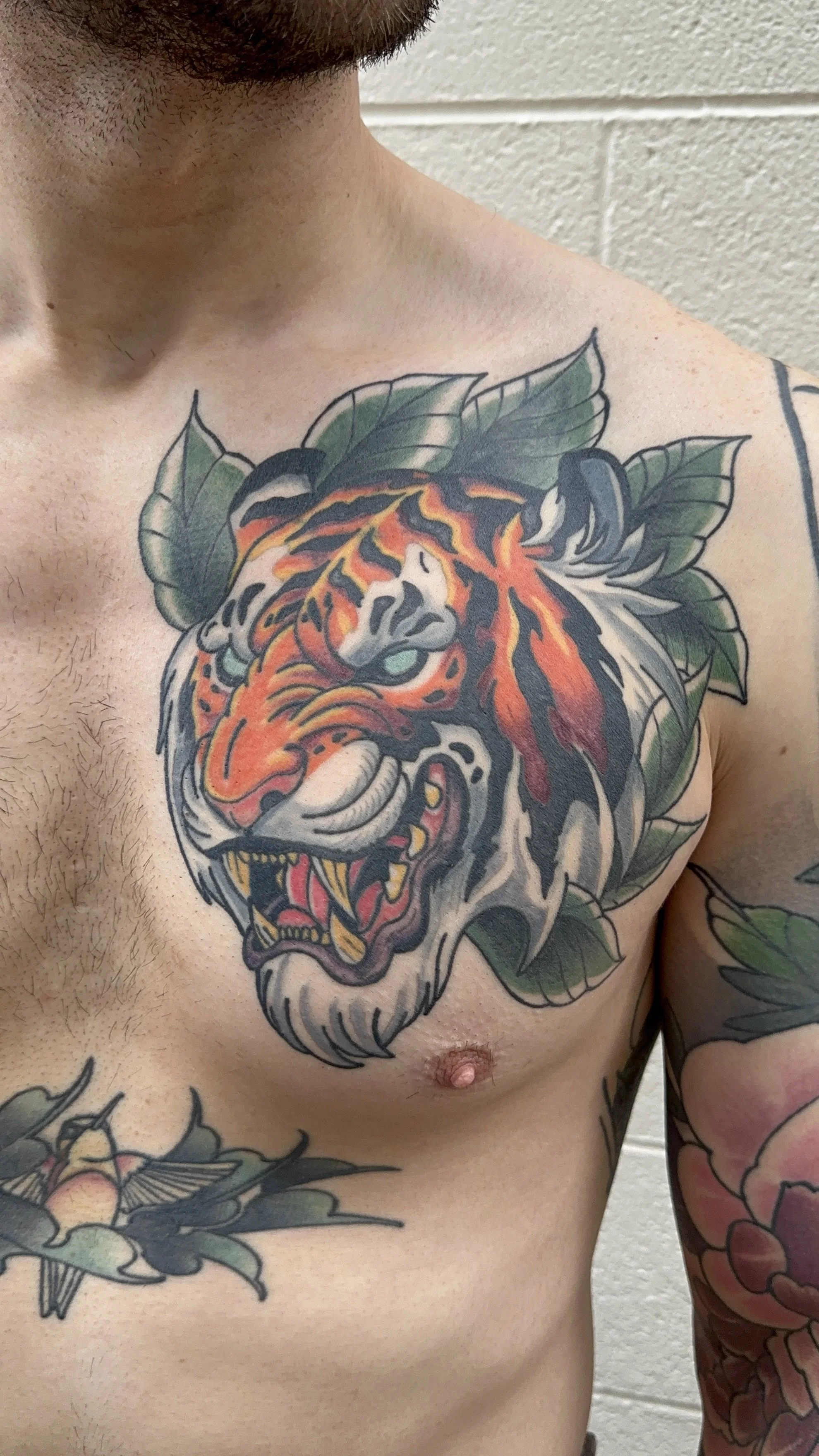 Tattoo of a fierce tiger head with orange, black, and white coloring, surrounded by green leaves on a man's chest.