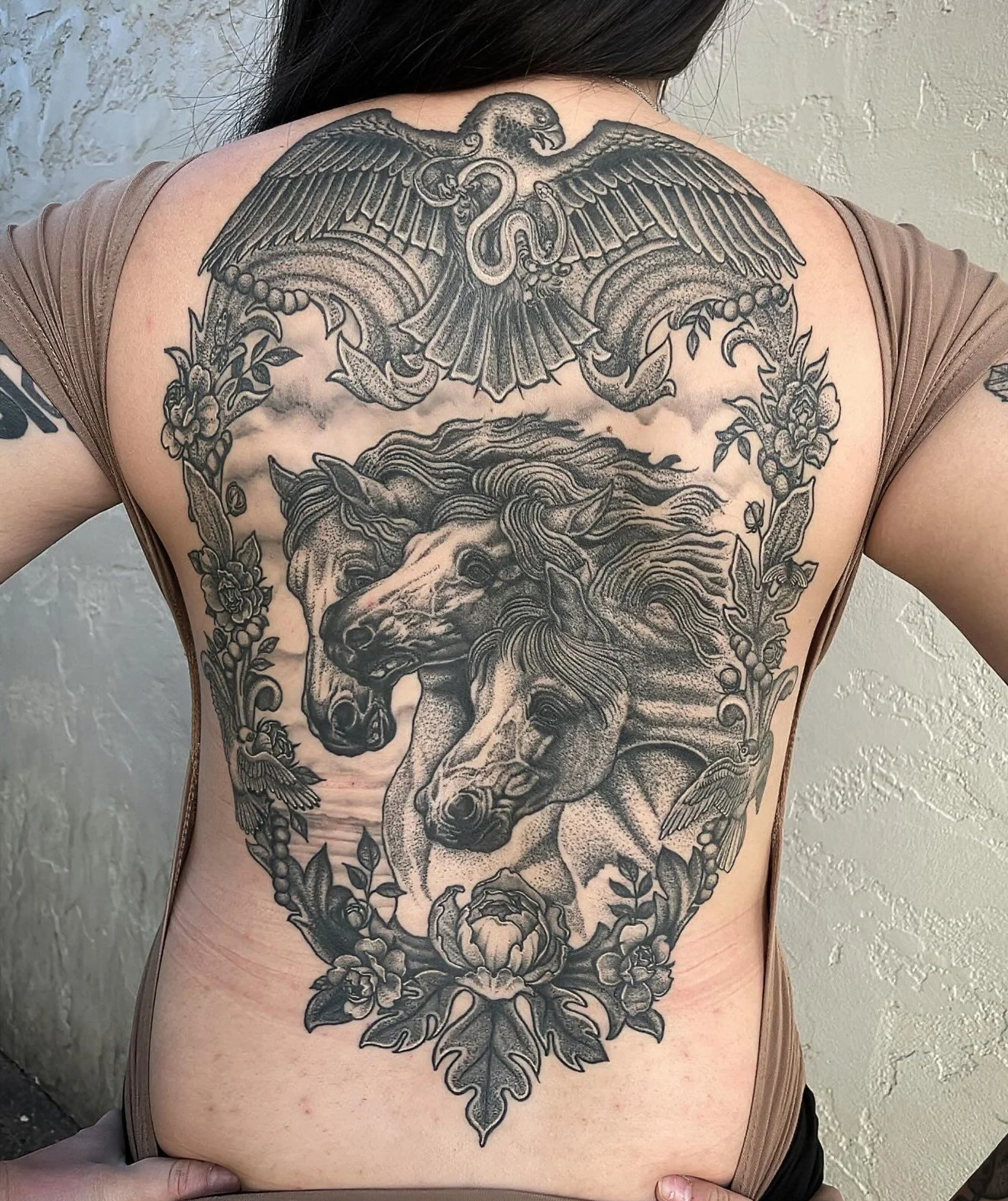 A large black and gray tattoo on a person's back depicting three horses' heads, surrounded by floral and decorative elements, and a pair of stylized birds above the horses.