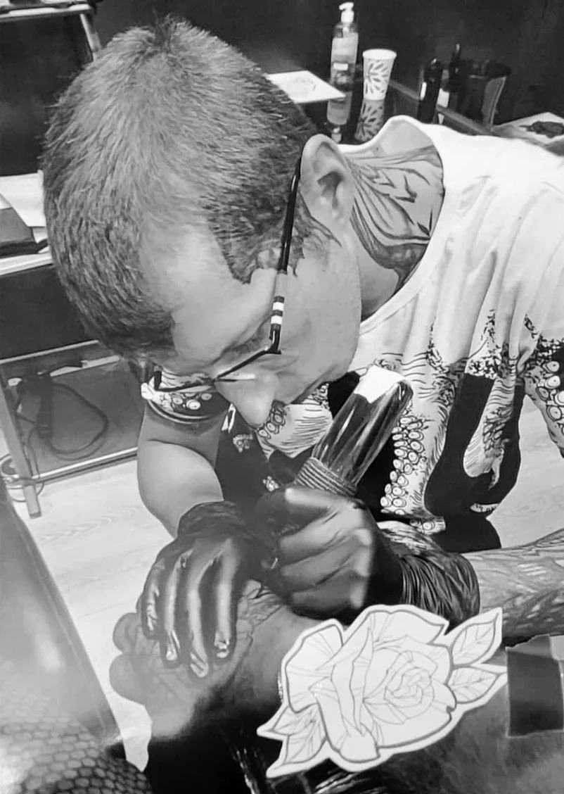 A person with glasses, tattoos, and short hair tattooing a hand with a tattoo machine in a tattoo studio. There is tattoo equipment and supplies on the counter in the background.