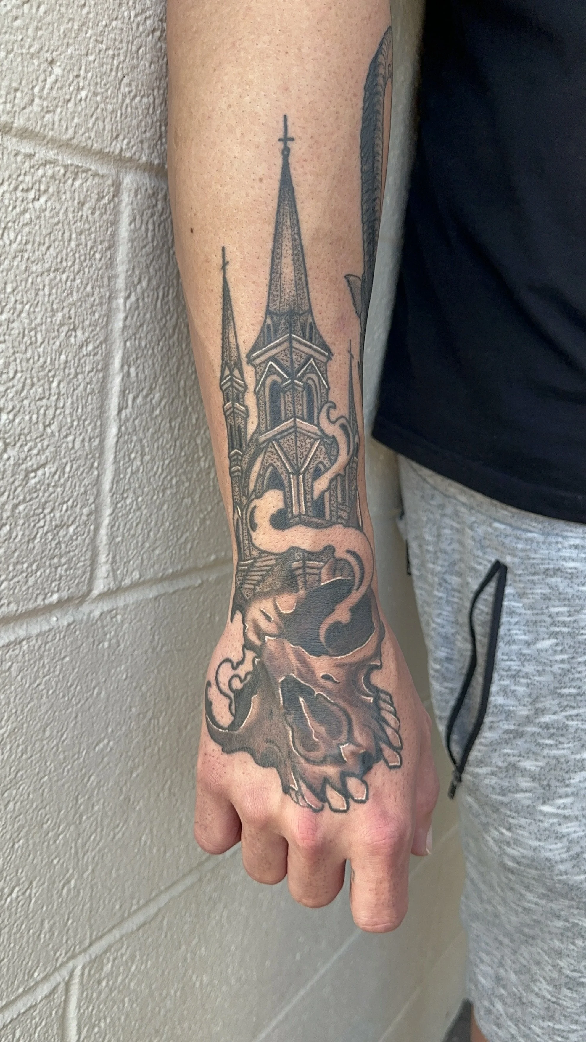 Tattoo of a church steeple with two smaller spires and a skull with smoke or flames rising from it on a person's forearm.