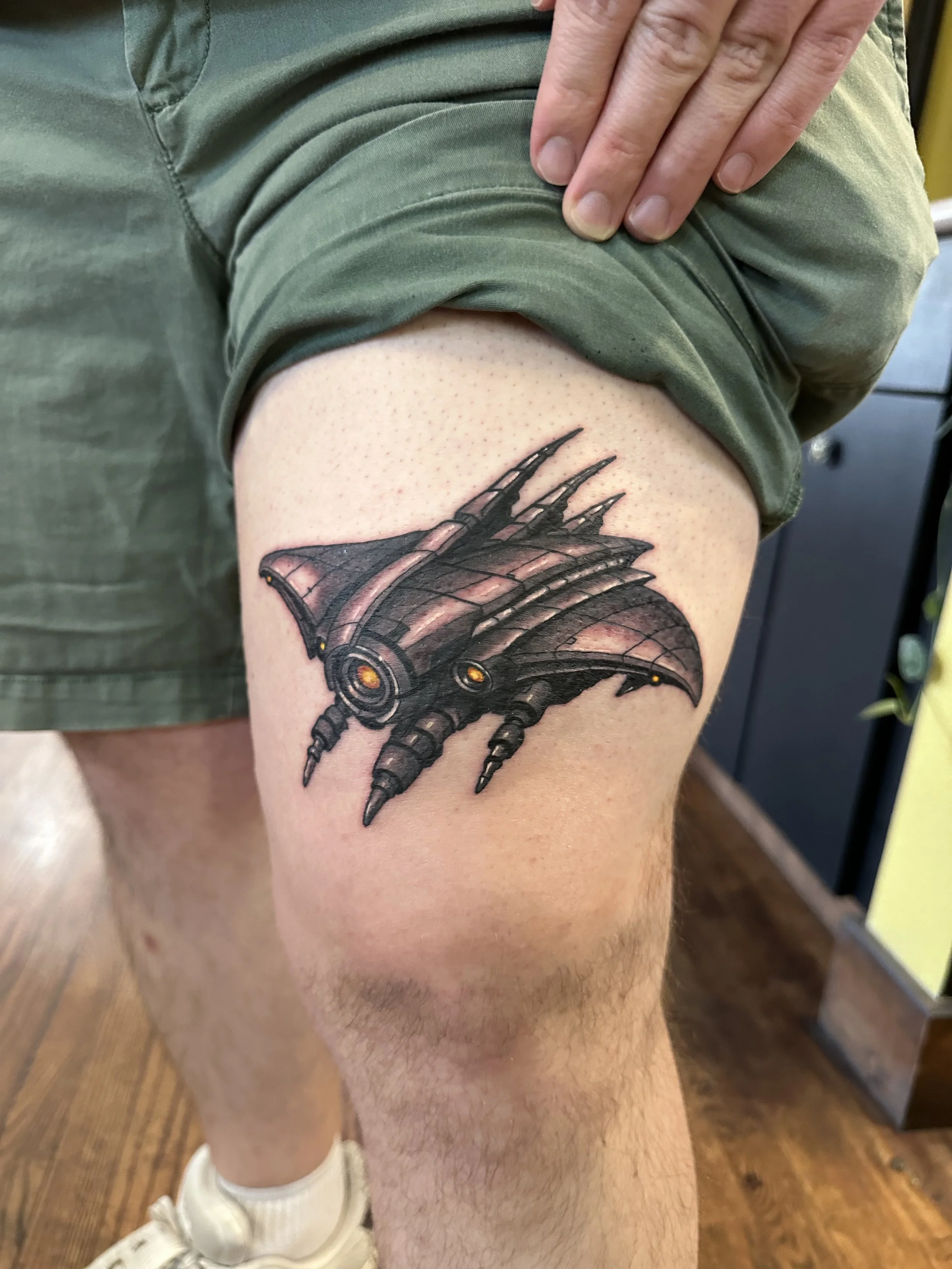 A person's upper thigh tattoo featuring a mechanical dragon wing design with detailed shading.