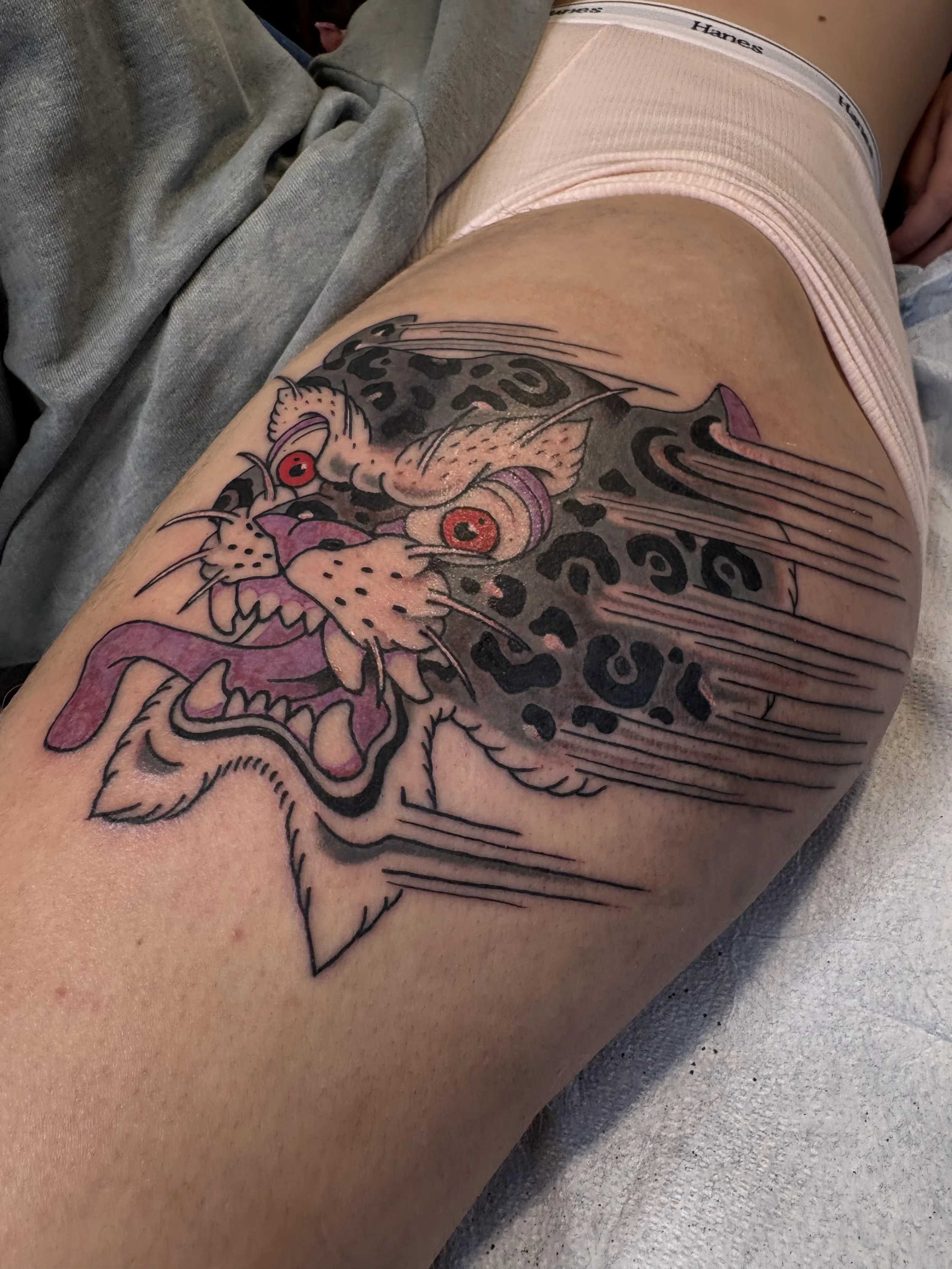 Tattoo of a colorful leopard's head with red eyes and purple accents on a person's thigh.