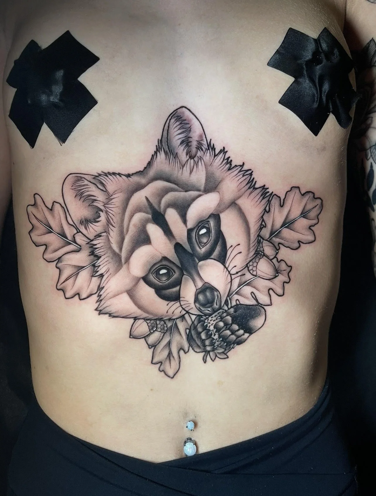 Tattoo of a raccoon face with leaves on the chest, black ribbons taped over the shoulders.