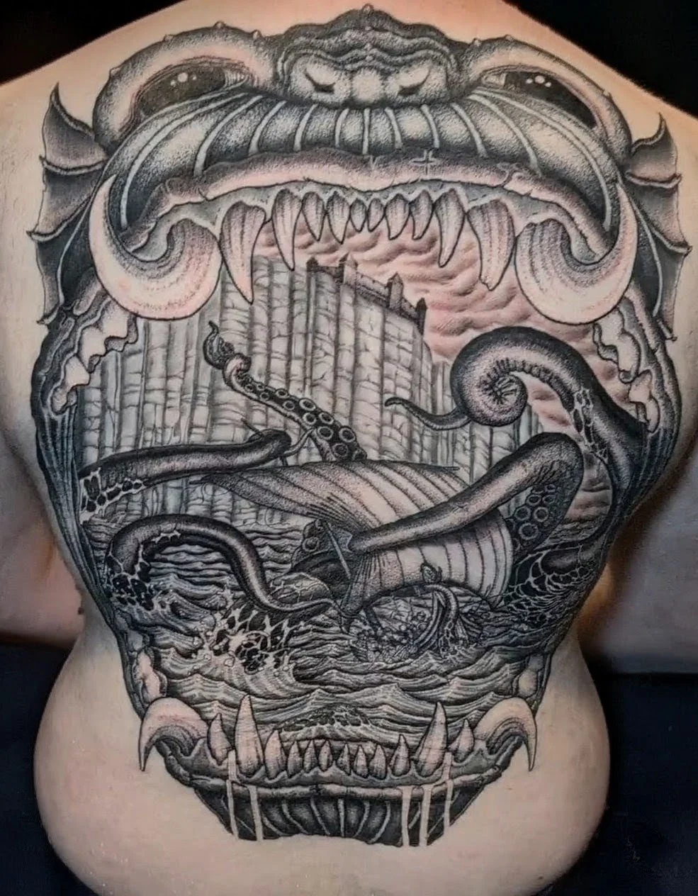 Black and gray tattoo on person's back depicting a monstrous creature's face with sharp teeth and fangs, tentacles, and a scene of an ocean with sharks and a large structure resembling a fortress or wall beneath a menacing sky.