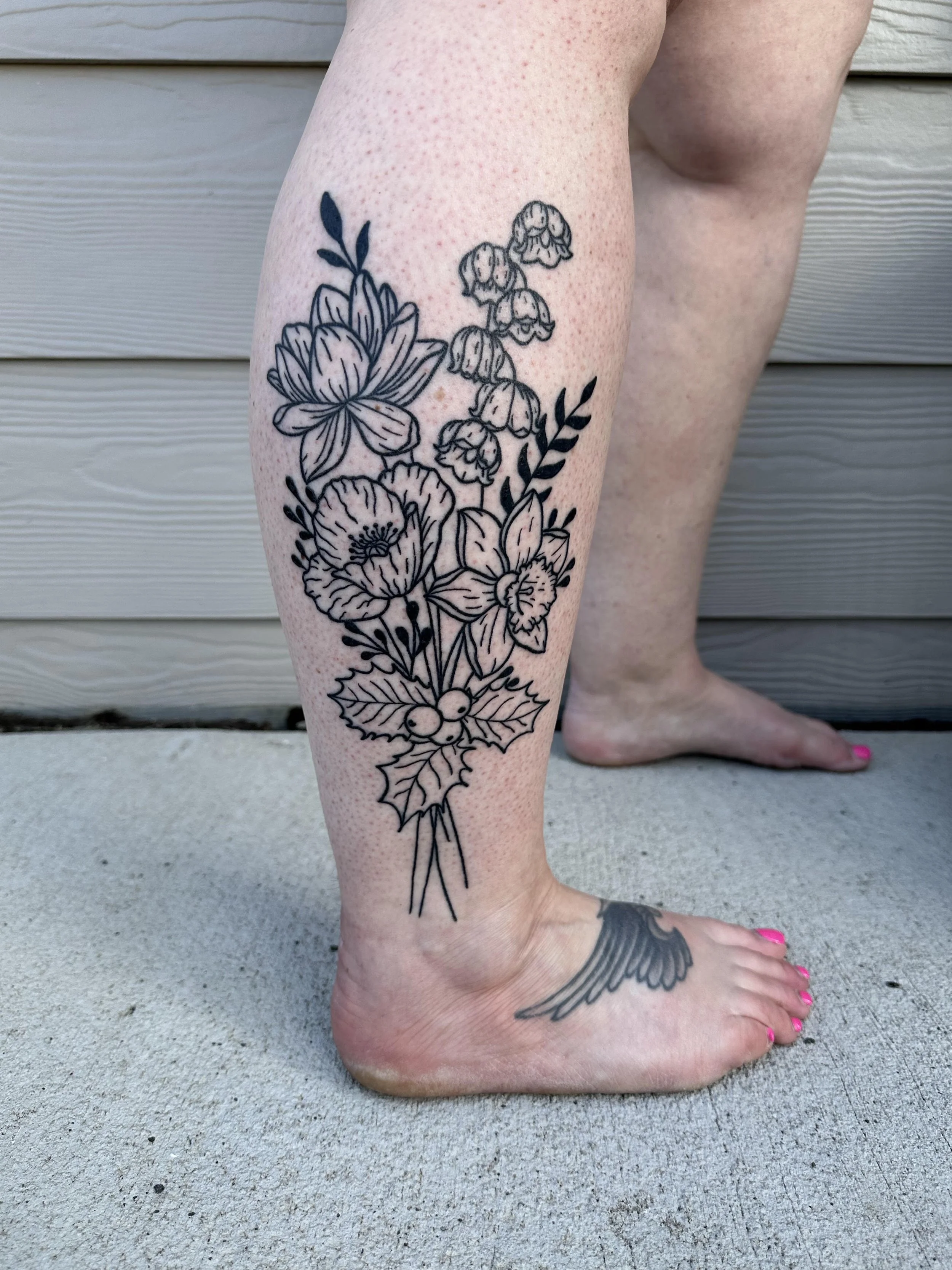 Black floral tattoo on person's calf and ankle, featuring holly leaves and berries, with a second tattoo of a bird's wing on the person's foot.